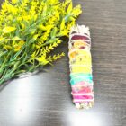 A 4" California White Sage Smudge Stick, infused with 7 Chakra colors, rests gracefully beside a stunning yellow flower.