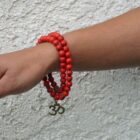 A woman's hand is holding a Red Howlite Gemstone Bead Double Om Charm Bracelet. The bracelet features a brilliant red howlite gemstone bead and is engraved, making it a personalized gemstone bracelet.