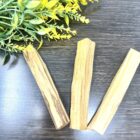 Three Palo Santo sticks - Thick on a table next to yellow flowers, providing Palo Santo Sticks - Thick | Home Fragrance| Healing| Spiritual home fragrance.