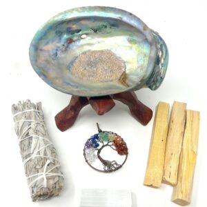 A Sage Smudge Kit with 4" California Organic White Sage, 3 Palo Santo Sticks, Abalone Shell, Wooden Tripod Stand, 7 Chakra Pendant & Selenite, displayed on a table.